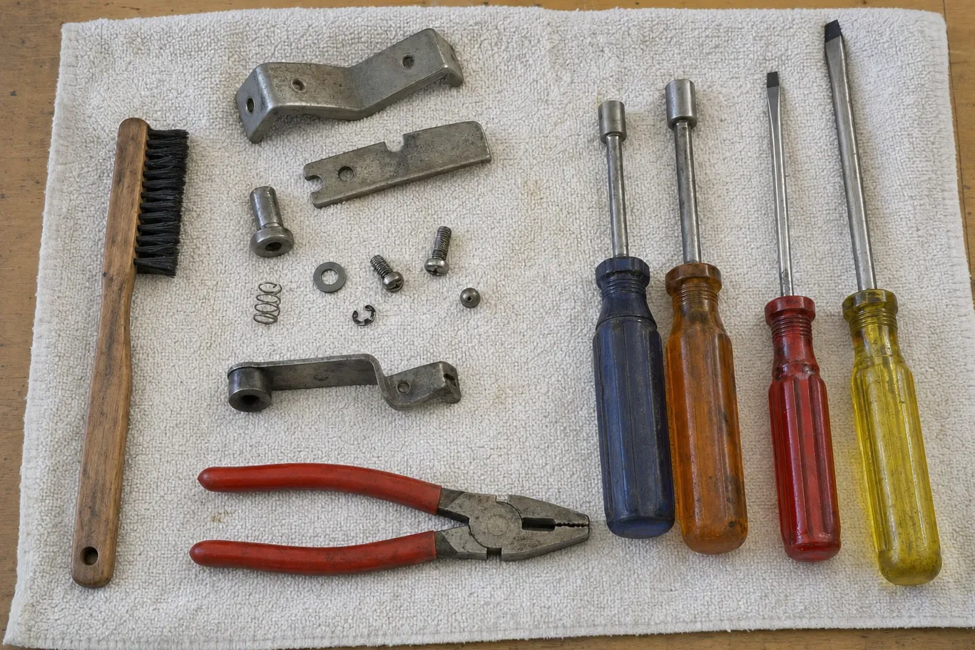 Wire brush, screwdrivers, pliers and a dismantled hinge and latch assembly with its screws and spring laid out on a towel